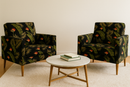 Two mid-century modern accent chairs upholstered in a dark tropical Birds of Paradise and palm leaf pattern, arranged around a round marble coffee table on a neutral rug.