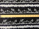 A close-up view of the Lagoon Nights Rayon fabric, displaying a repeating tropical pattern of white palm trees, tiki huts, and sailboats on a dark navy blue background. A wooden ruler is placed horizontally across the center of the fabric to indicate scale, showing that the repeating horizontal bands of the island scenery are approximately 3 to 4 inches in height.