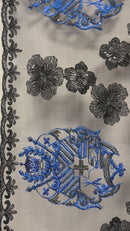 Close-up of a "Black & Blue" 100% polyester lace fabric featuring an intricate embroidered blue and gold Tongan seal (National Coat of Arms) alongside black sequined hibiscus flowers and a decorative black lace border on a sheer off-white mesh.